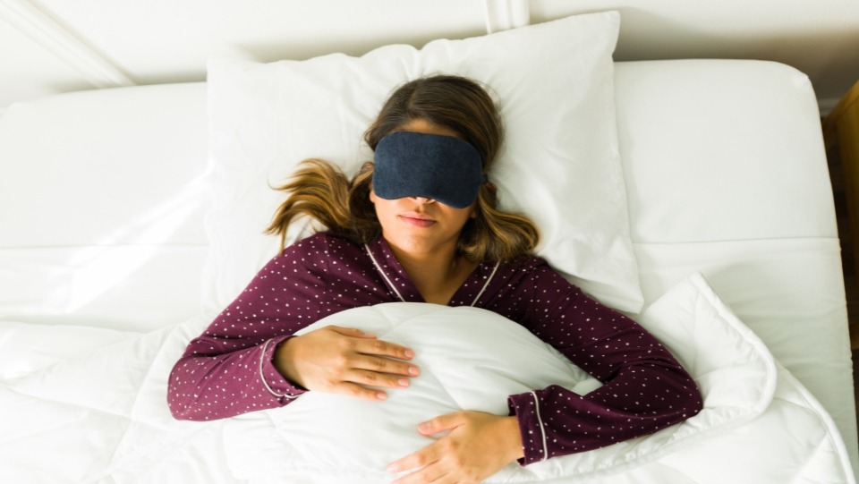 A woman lying in bed wearing a sleep mask, experiencing the benefits of using sleep masks for better sleep quality and brain function, as she embraces a pillow in a peaceful sleeping environment.